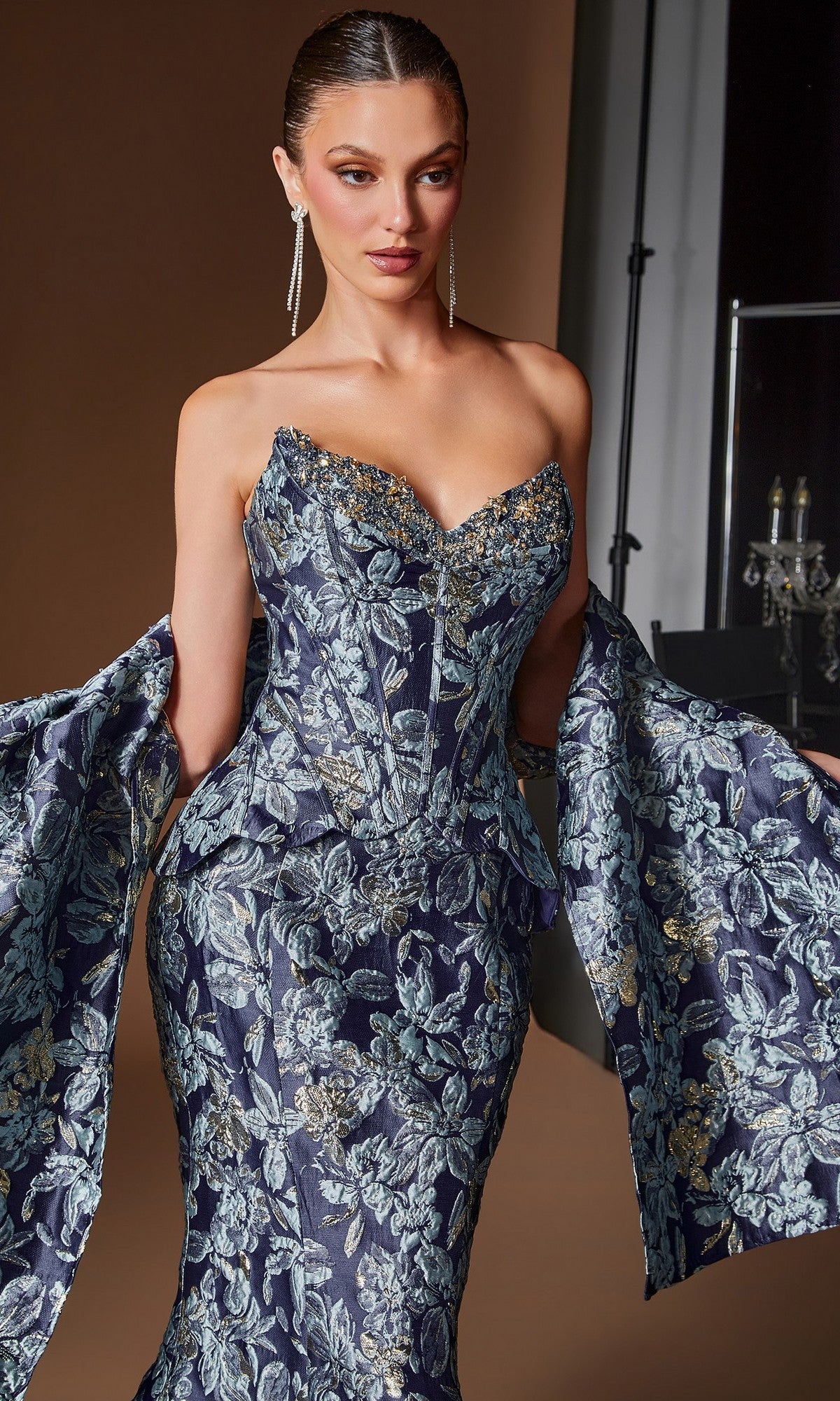 A woman poses against a neutral background wearing the Ladivine CD655 long formal dress, an ornate strapless blue and silver floral jacquard evening gown featuring a corset bodice and dramatic sleeves.